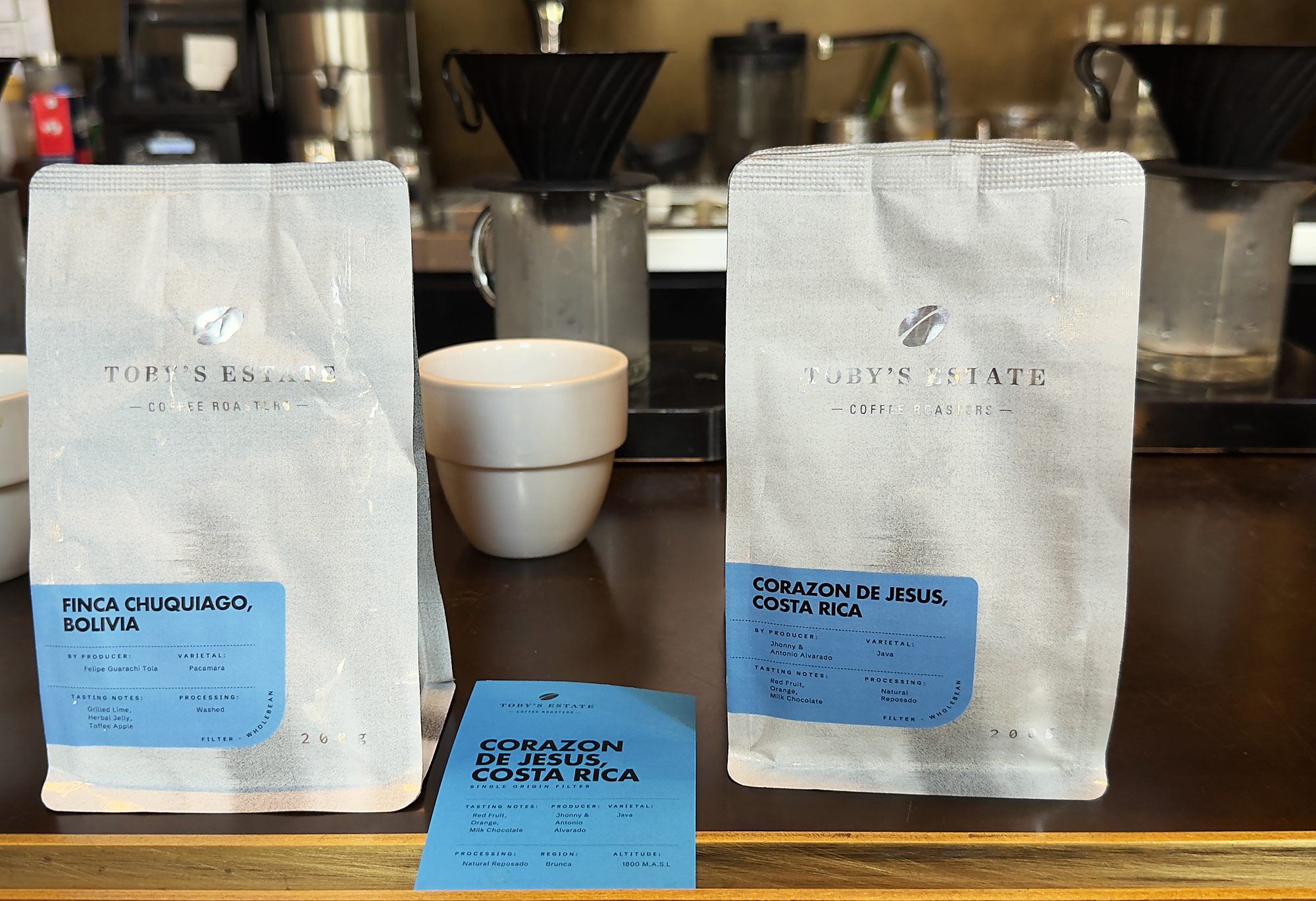Two coffee bags standing on a cafe bar with a cup between them, Finca Chuquiago Bolivia and Corazon de Jesus Costa Rica, and a printed lot card in front