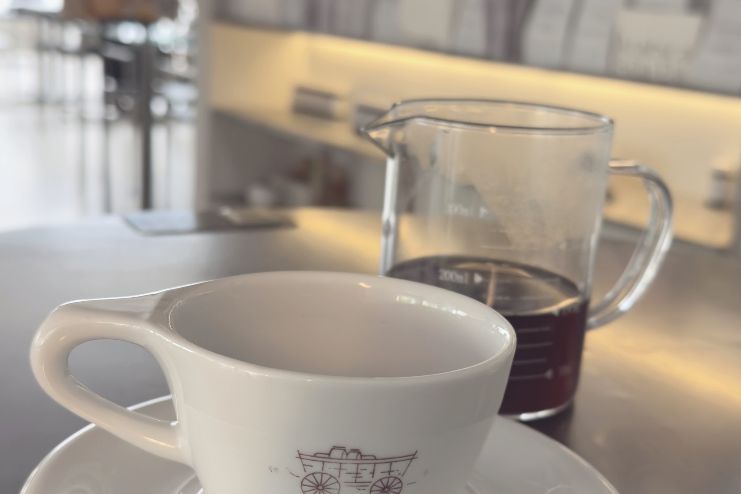 A cup and glass carafe on a counter, with a wall of coffee bags behind